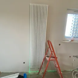 Biały, pionowy grzejnik panelowy zamontowany na ścianie, wyznaczony poziom laserem krzyżowym, obok czerwona drabina, widoczne ślady po instalacji hydraulicznej w ścianie.