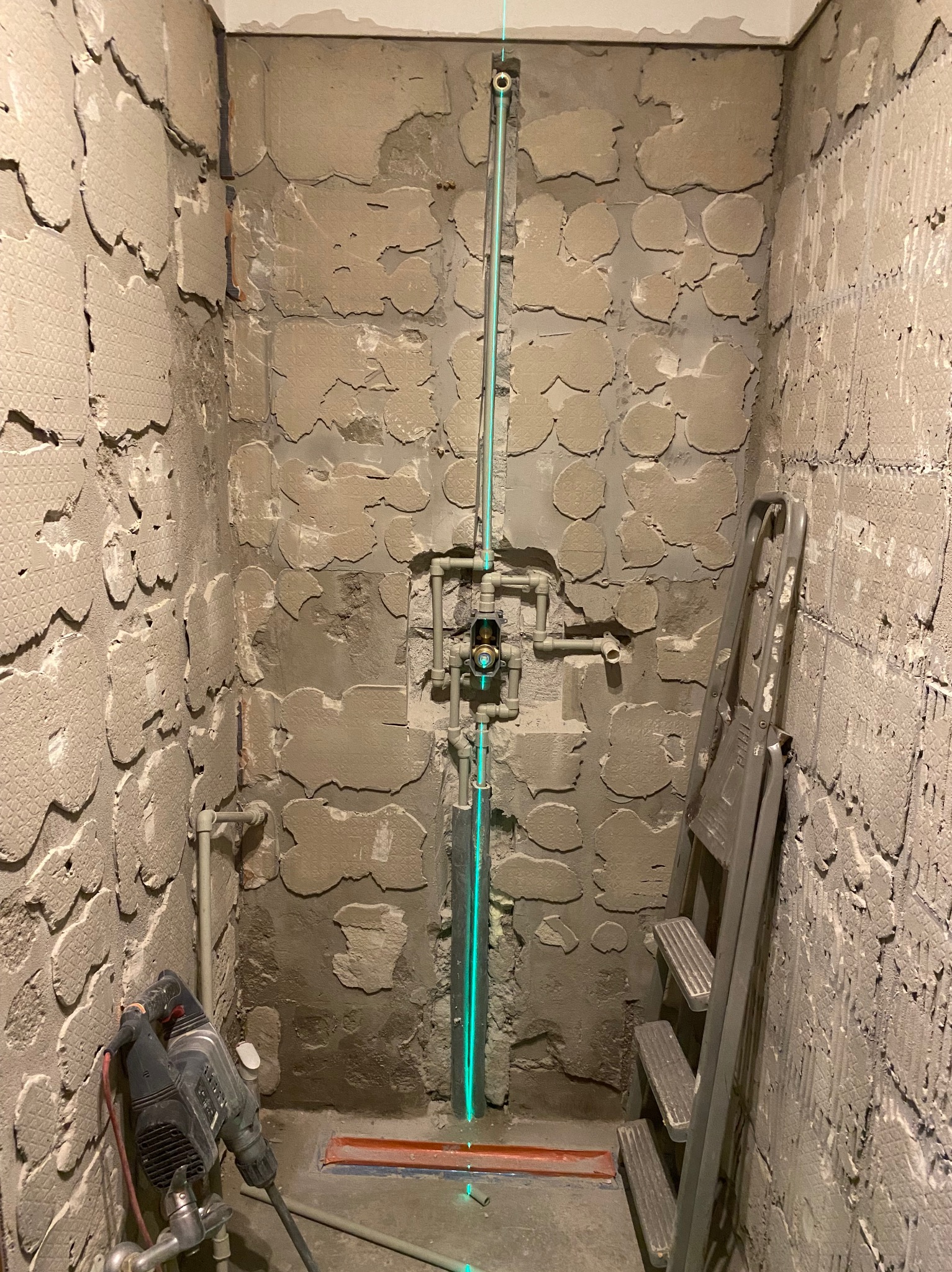 Surowe ściany łazienki w trakcie remontu, widoczna instalacja wodna z zielonym laserem poziomującym, drabina i młot udarowy.