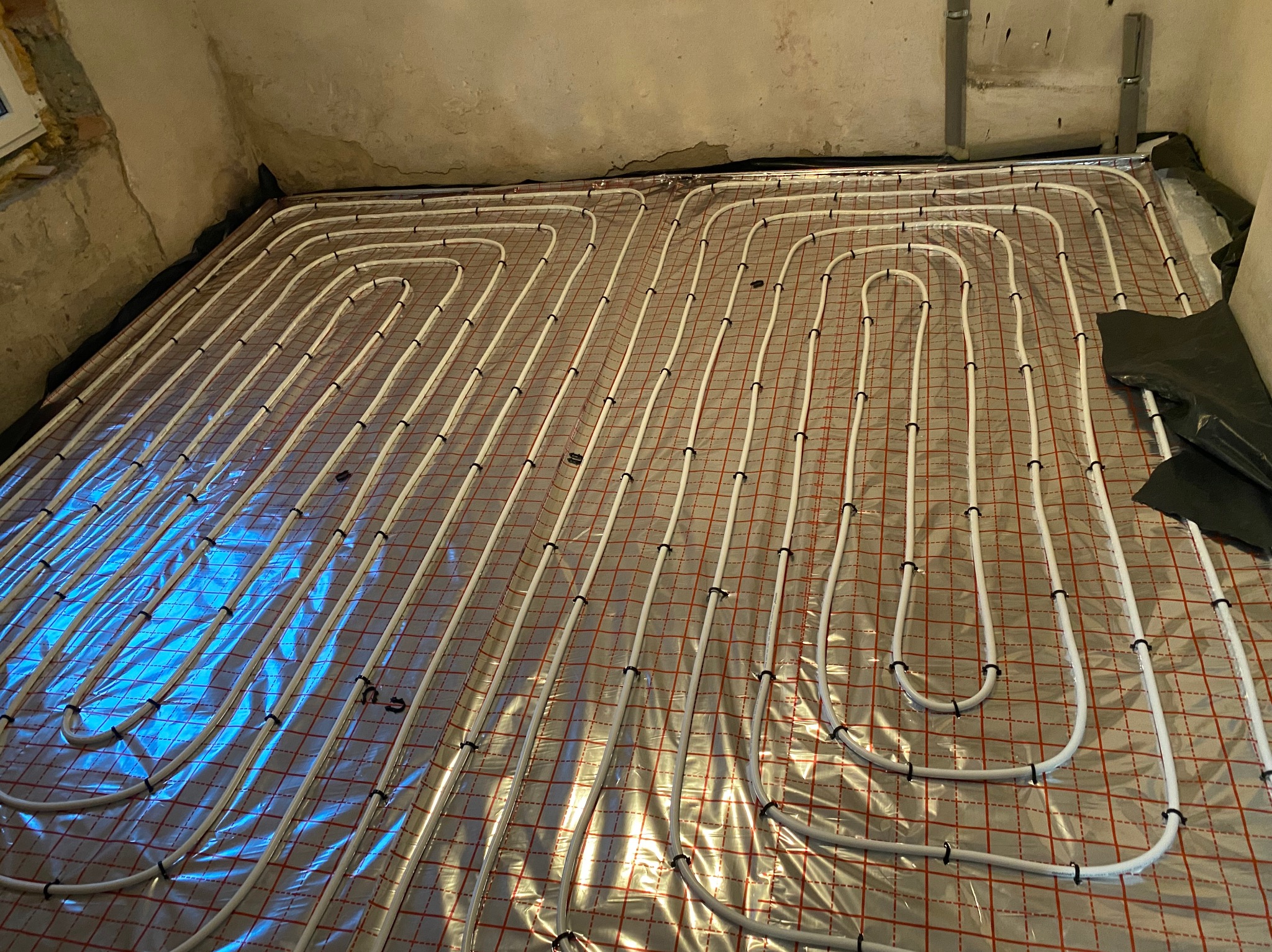 Białe rury grzewcze ułożone w pętle na folii z czerwoną kratką, montowane do podłogowego systemu grzewczego w surowym pomieszczeniu.