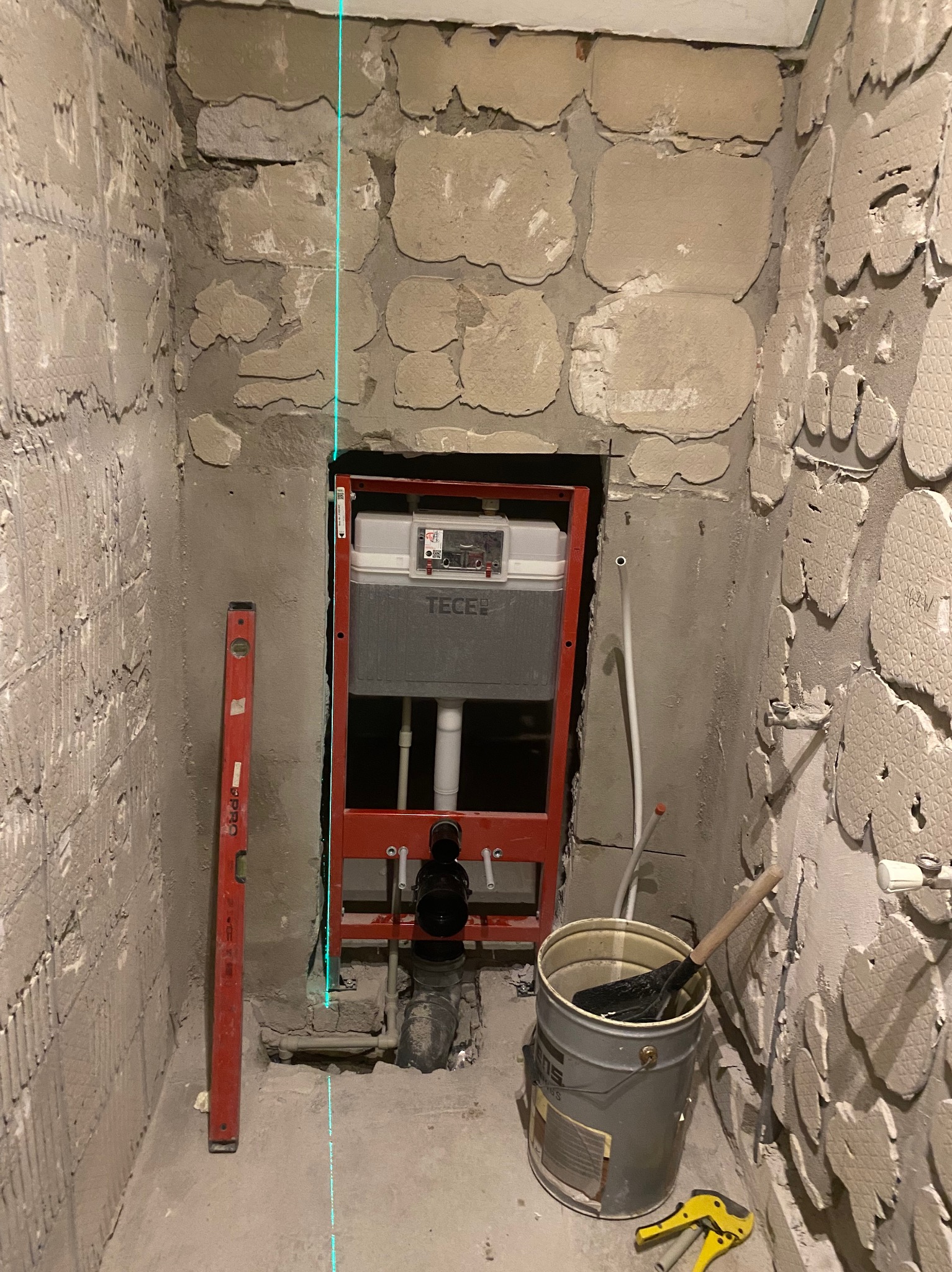 Montaż stelaża podtynkowego TECE w surowej łazience, widoczna poziomica laserowa, wiadro z narzędziami i fragment ściany z resztkami płytek.