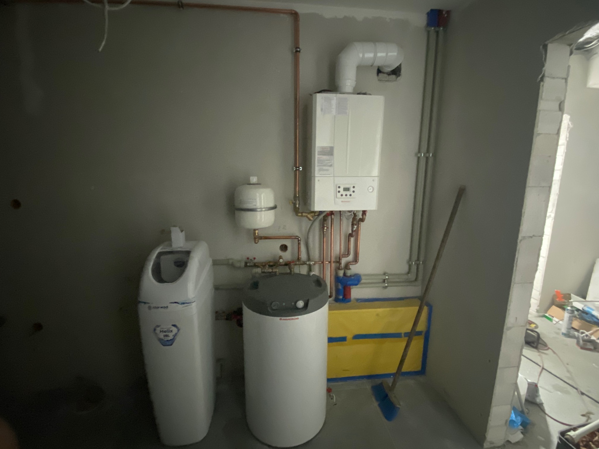 Nowa instalacja grzewcza w surowym wnętrzu: piec gazowy, zasobnik ciepłej wody, zmiękczacz wody, rury miedziane i plastikowe, żółta obudowa, ściany w stanie deweloperskim.
