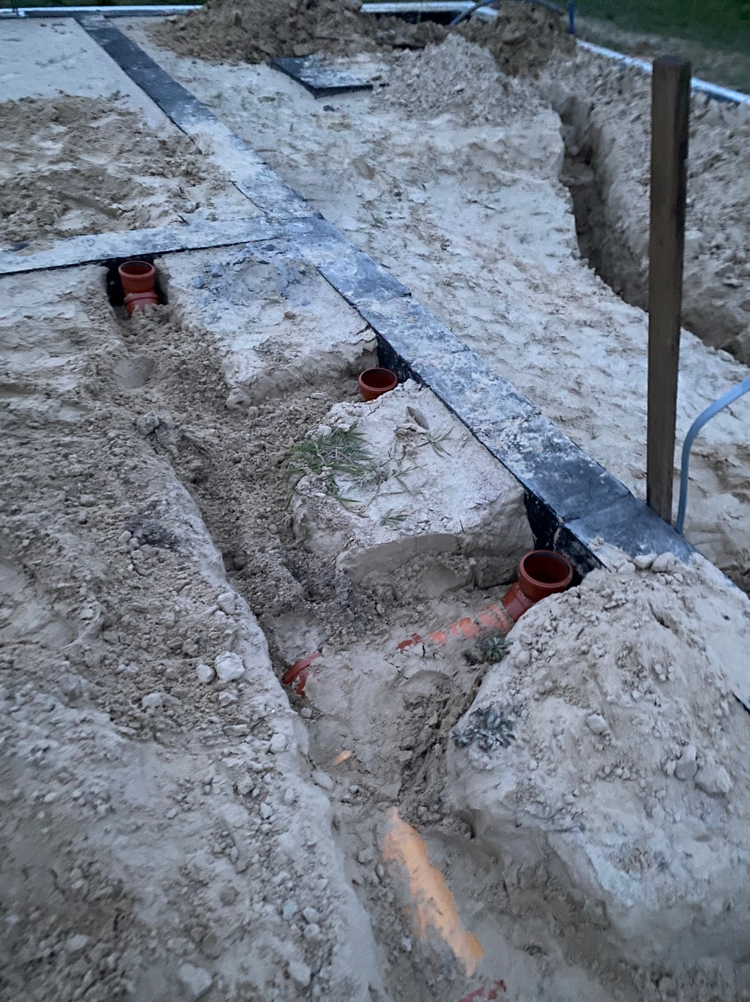 Wykopy w piasku z widocznymi rurami instalacji podposadzkowej i czarną taśmą wyznaczającą przebieg instalacji.