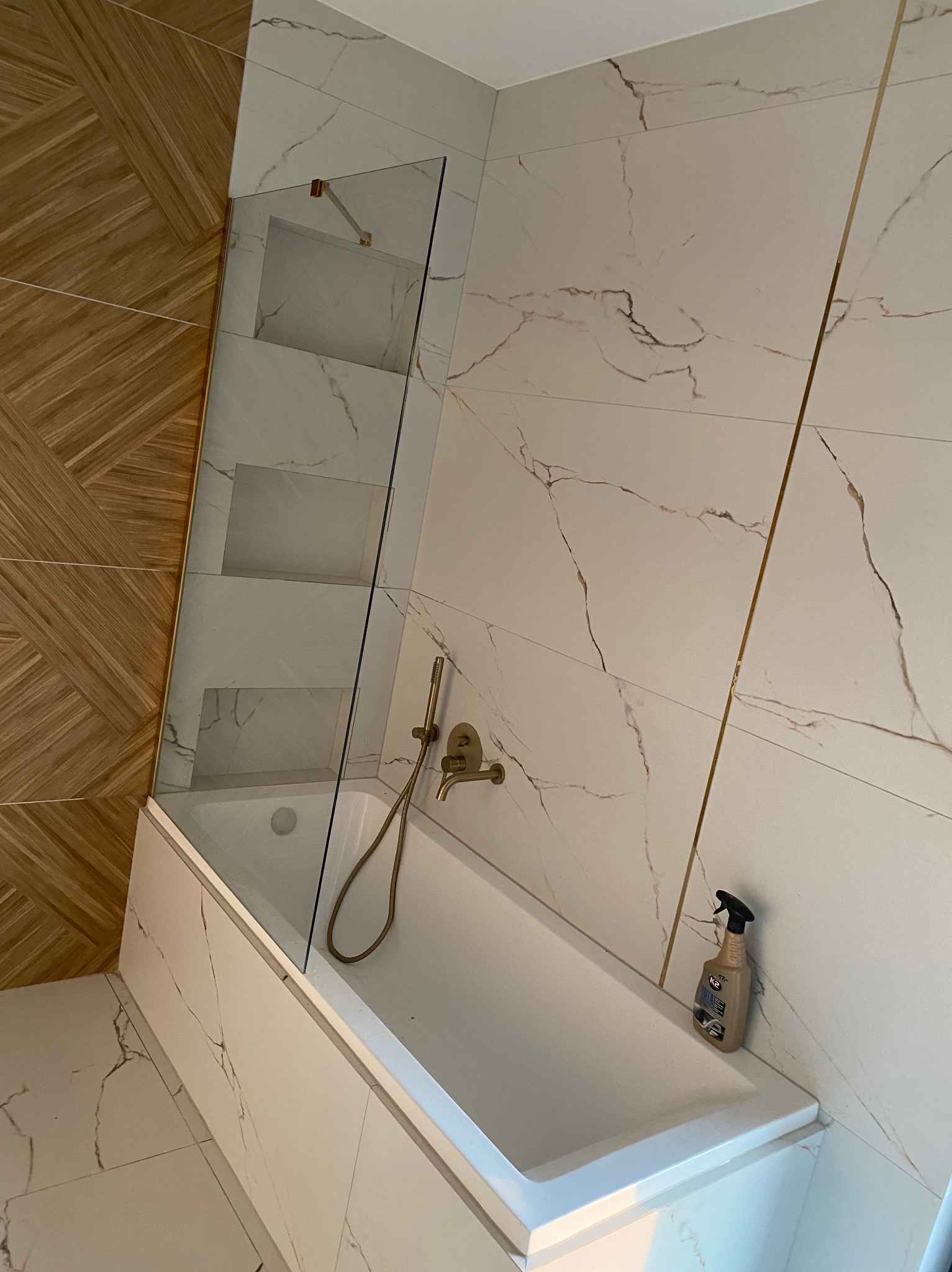 Biała wanna z zamontowanym szklanym parawanem, bateria wannowa w kolorze złotym i butelka płynu do czyszczenia na brzegu wanny, wykończenie ścian płytkami imitującymi marmur z złotymi wstawkami...