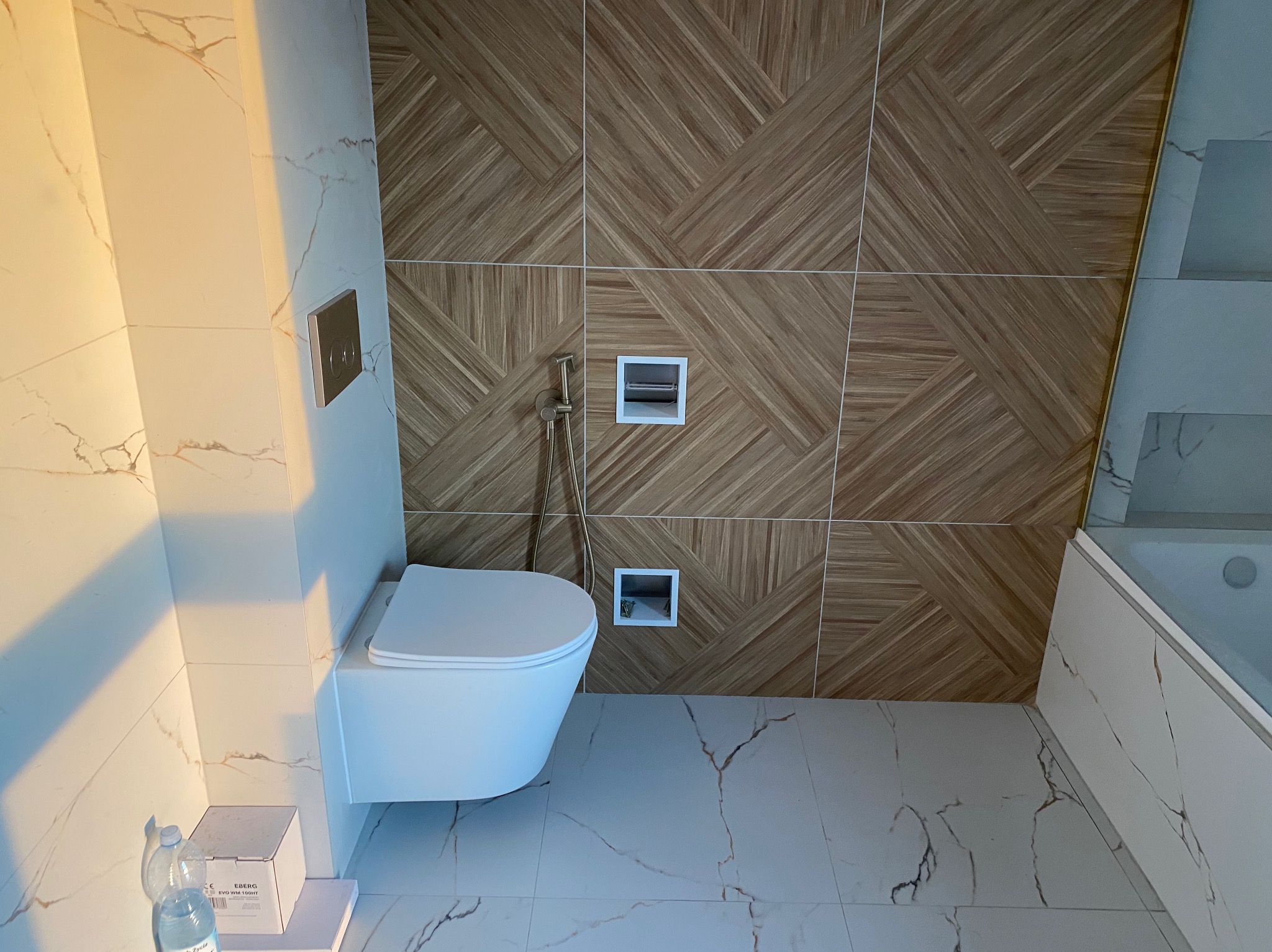 Nowoczesna toaleta podwieszana z chromowanym przyciskiem spłukującym i bidetką w łazience wykończonej płytkami imitującymi marmur i drewno.