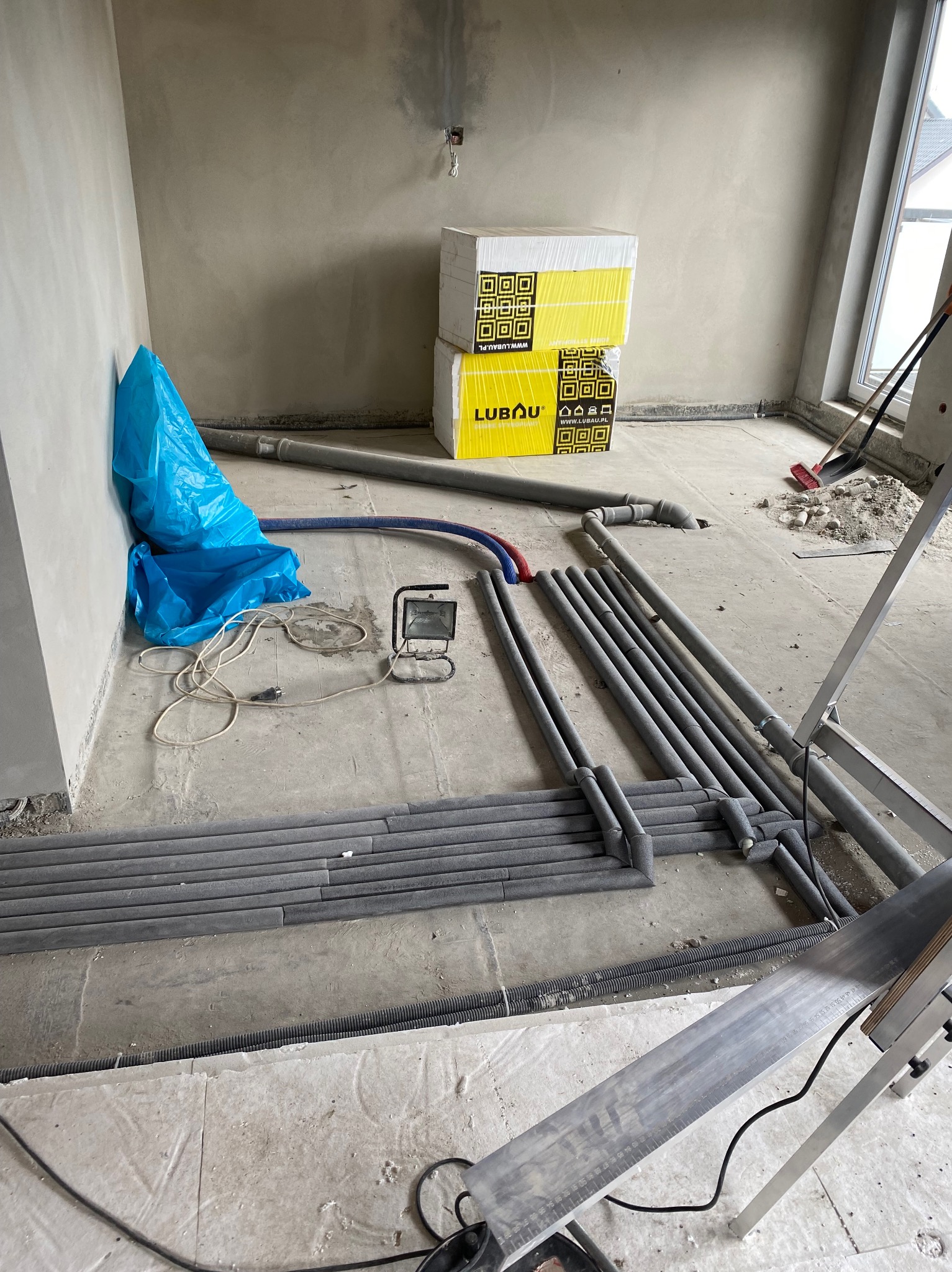 Niedokończona instalacja w surowym wnętrzu: rury w izolacji, niebieski worek, lampa halogenowa, opakowania materiałów budowlanych Lubau, widok z góry na elementy instalacji.