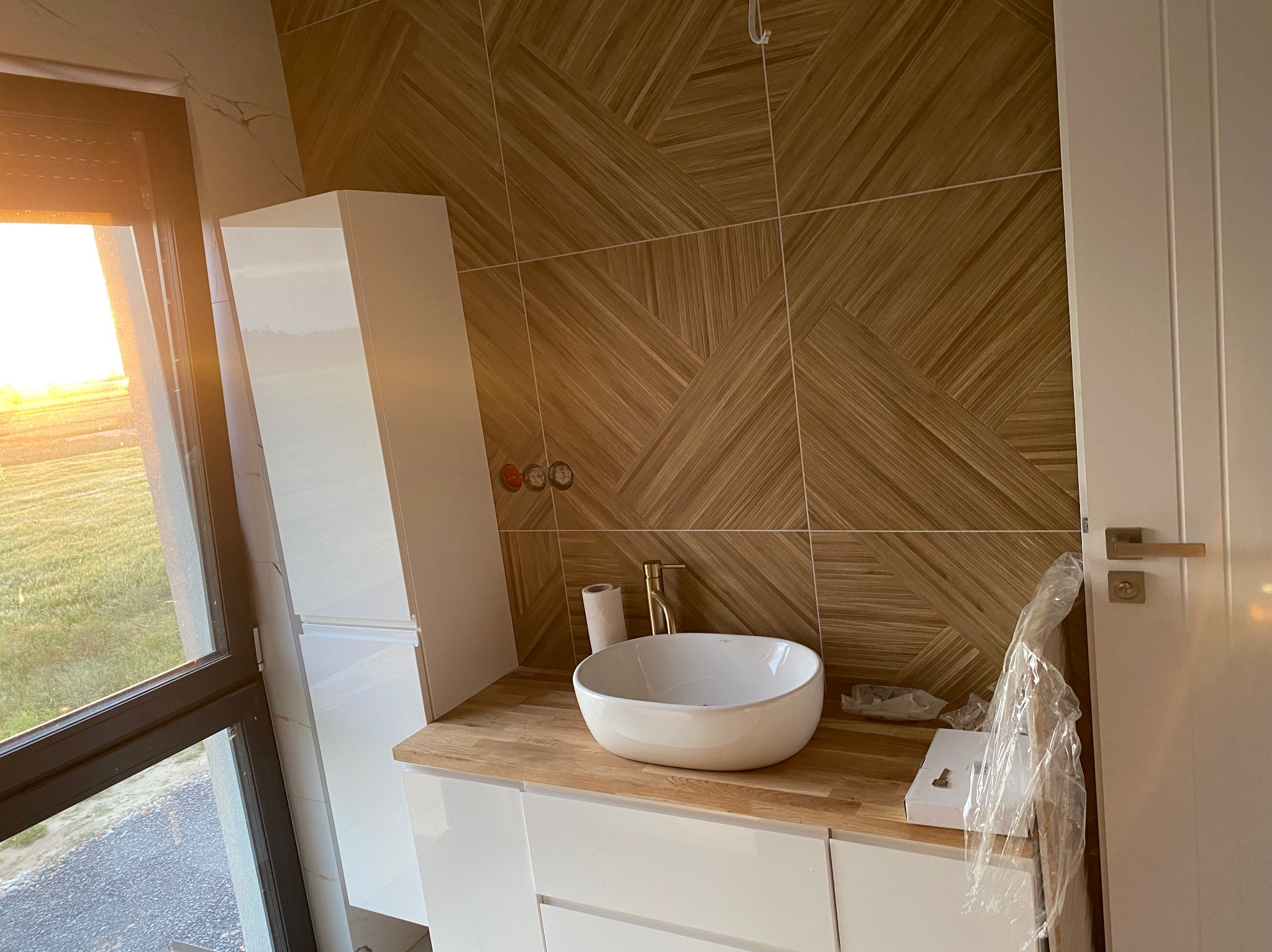 Nowoczesna łazienka w trakcie wykańczania: biała umywalka nablatowa, złota bateria, drewnopodobne płytki na ścianie ułożone w geometryczny wzór, widok na zachodzące słońce przez okno.