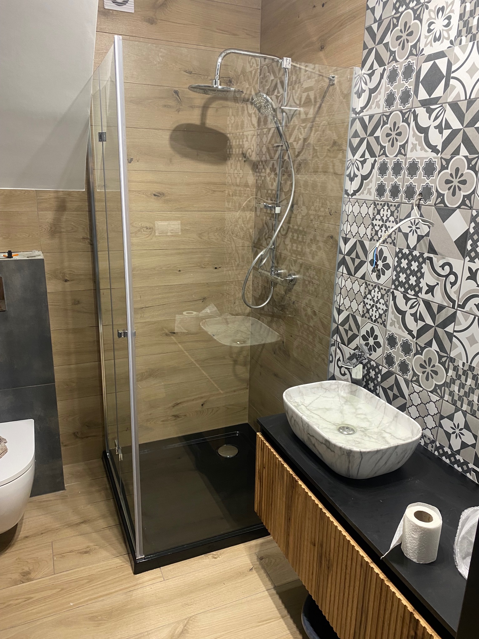 Nowoczesna łazienka z kabiną prysznicową typu walk-in z czarnym brodzikiem, marmurową umywalką nablatową i płytkami w geometryczne wzory na ścianie.