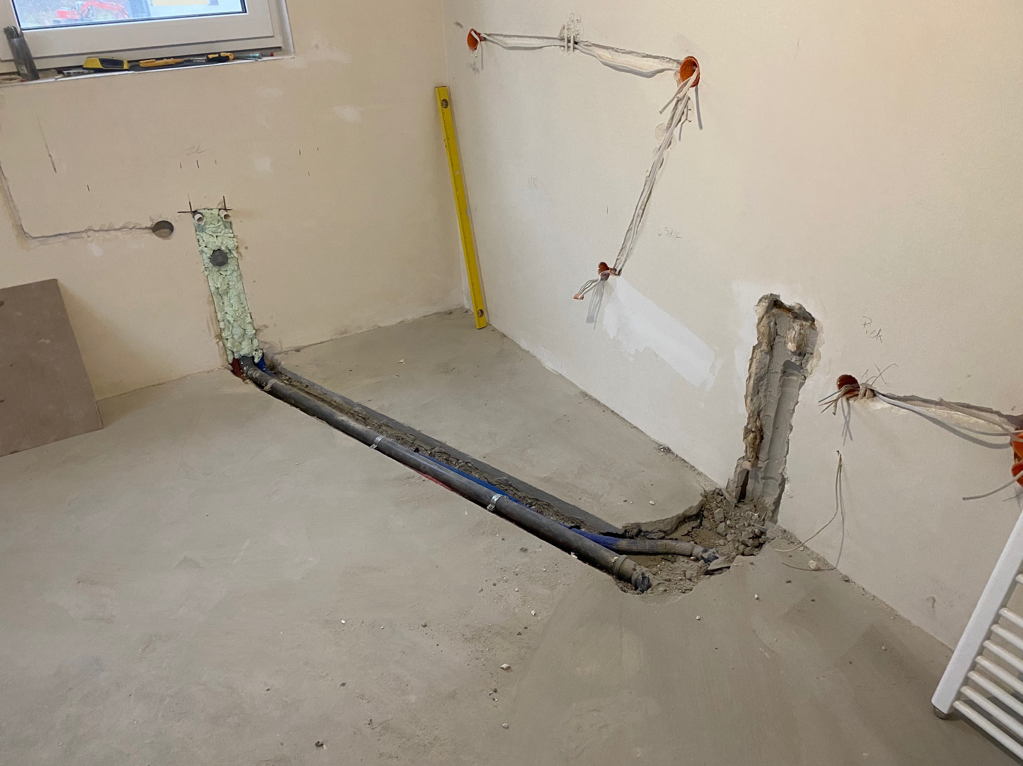 Przygotowanie podłoża pod instalację: rury grzewcze i wodne w wyciętych bruzdach w posadzce, zabezpieczone pianką montażową, widoczne wyprowadzenia elektryczne na ścianach i poziomica.