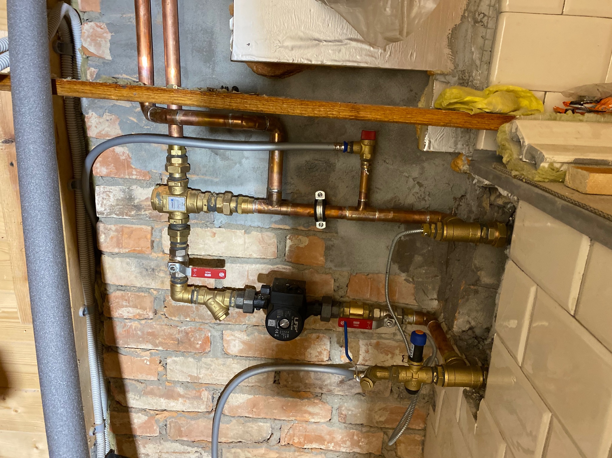 Fragment instalacji hydraulicznej z miedzianymi rurami, zaworami z czerwonymi i niebieskimi pokrętłami, pompą obiegową LFP, zamontowanej na tle ściany z czerwonej cegły i częściowo otynkowanej...