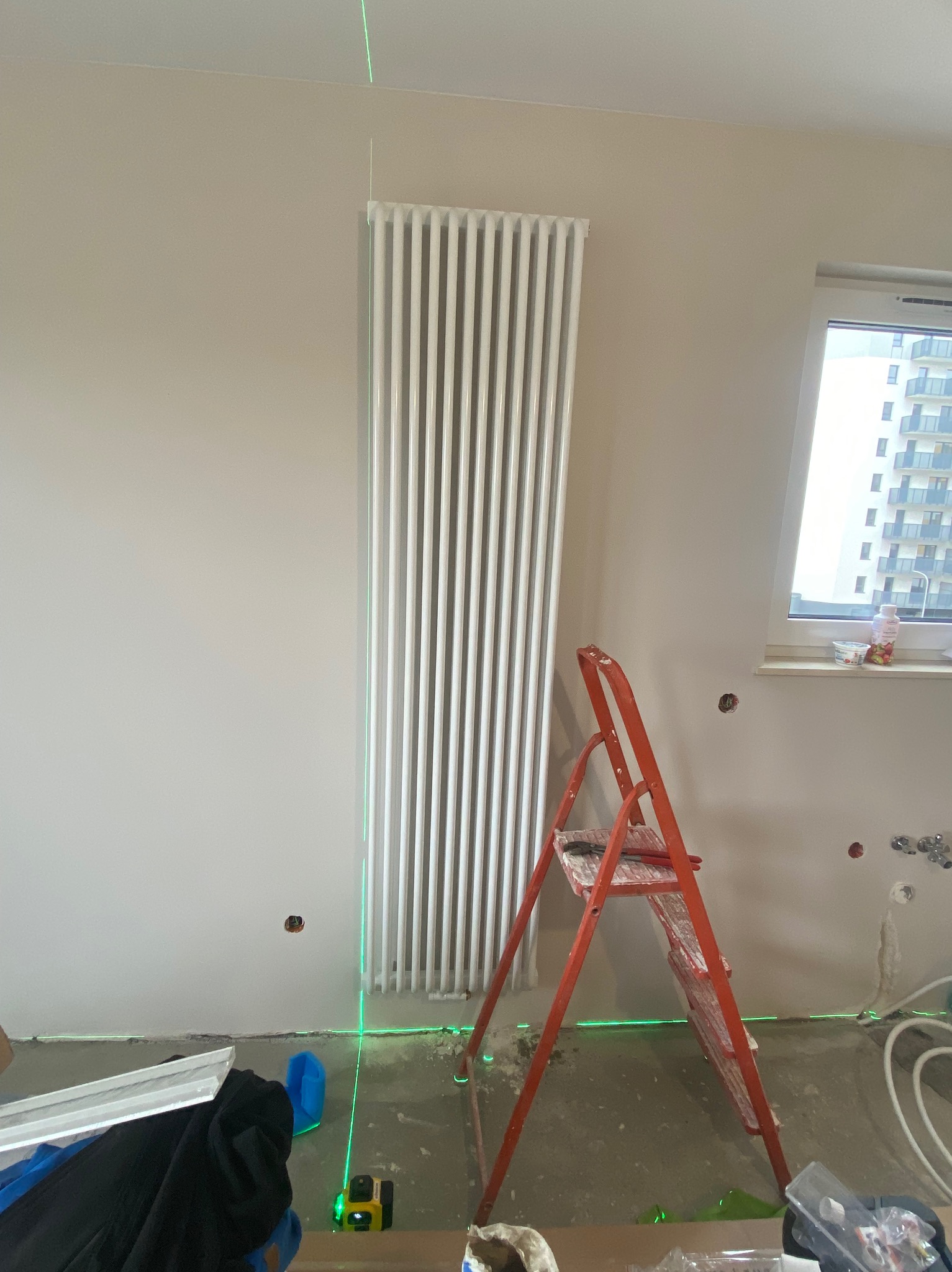 Biały, pionowy grzejnik panelowy zamontowany na ścianie, wyznaczony poziom laserem krzyżowym, obok czerwona drabina, widoczne ślady po instalacji hydraulicznej w ścianie.
