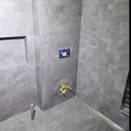 Wnętrze łazienki w trakcie prac wykończeniowych: szare płytki imitujące kamień na ścianach i podłodze, widoczne elementy instalacji podtynkowej, niezabudowany fragment sufitu z płyt...