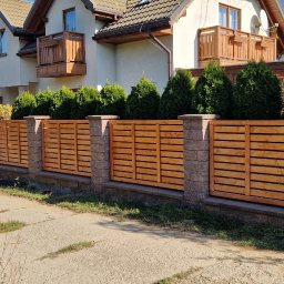 Meble & Tarasy - Nowoczesne ogrodzenie z poziomych desek w ciepłym odcieniu drewna, osadzone na kamiennych słupkach i podmurówce, na tle dwupiętrowego domu z balkonami i zadbanym żywopłotem.