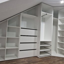 Meble & Tarasy - Biała zabudowa meblowa pod skosem dachu, z otwartymi półkami i szufladami z czarnymi akcentami, na drewnianej podłodze.