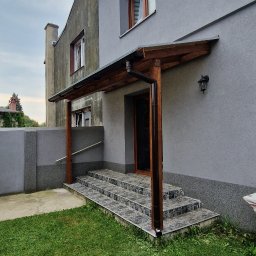 Meble & Tarasy - Zadaszone wejście do domu z drewnianą konstrukcją wspartą na słupach, prowadzące po schodach wyłożonych płytkami, z widocznym trawnikiem i fragmentem elewacji budynku.
