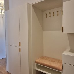 Meble & Tarasy - Biała zabudowa przedpokoju z siedziskiem obitym pluszem w odcieniu brzoskwini i złotymi haczykami na ścianie, widoczny fragment drewnianej podłogi oraz podłogi imitującej marmur.