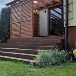 Drewniany taras z zadaszeniem z poliwęglanu, osłonięty panelami z poziomych desek, schody z desek prowadzą na trawnik.