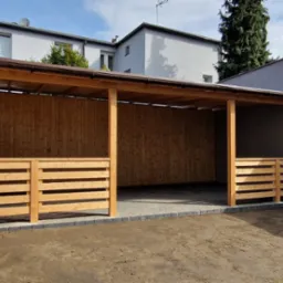 Drewniana konstrukcja z zadaszeniem, częściowo osłonięta, z niskimi płotkami, na świeżo wyłożonym bruku, otoczona ziemią i budynkami.