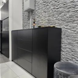 Czarna komoda z szufladami i szafkami na tle ściany wyłożonej szarym kamieniem dekoracyjnym, obok pufa w kolorze grafitowym.