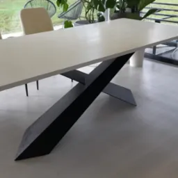 Stół z białym blatem i czarną, geometryczną podstawą w kształcie litery X, otoczony krzesłami w jasnych kolorach, na tle okna z widokiem na zieleń.