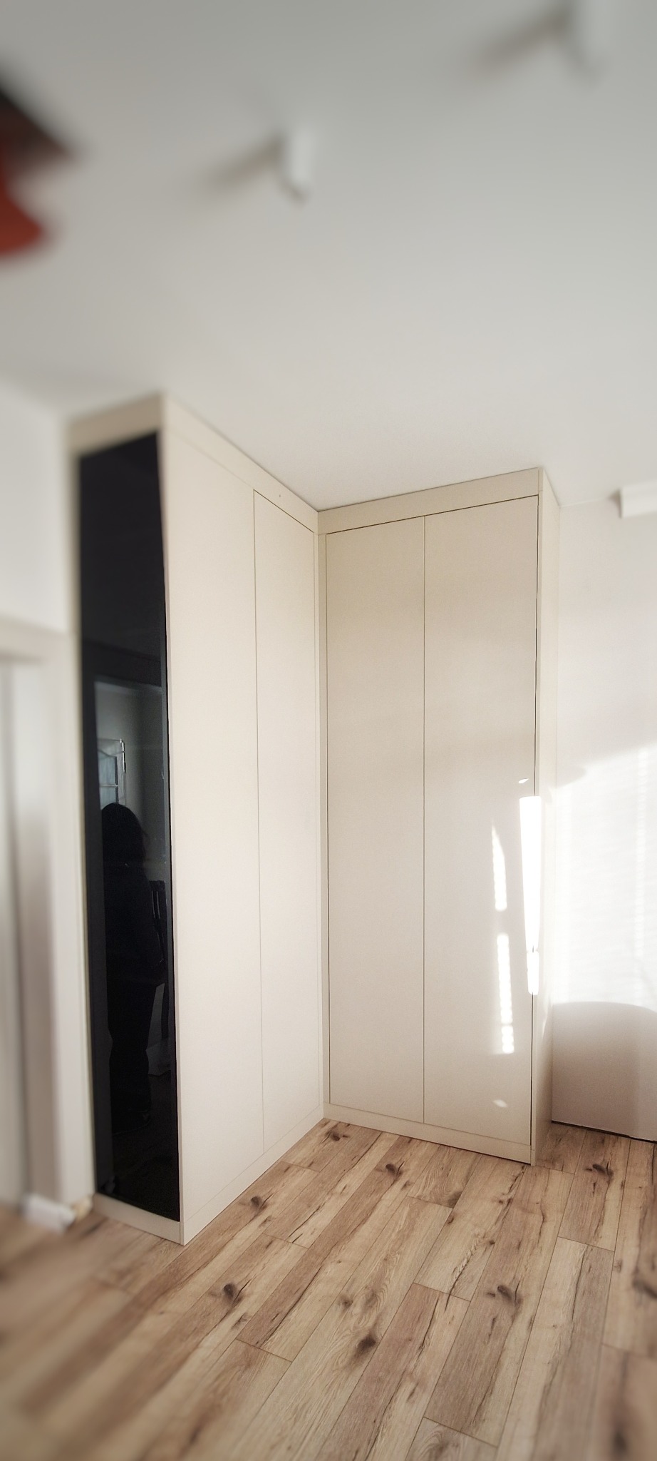 Jasna, narożna zabudowa meblowa z czarnym, lustrzanym frontem, drewniana podłoga, minimalistyczne wnętrze.