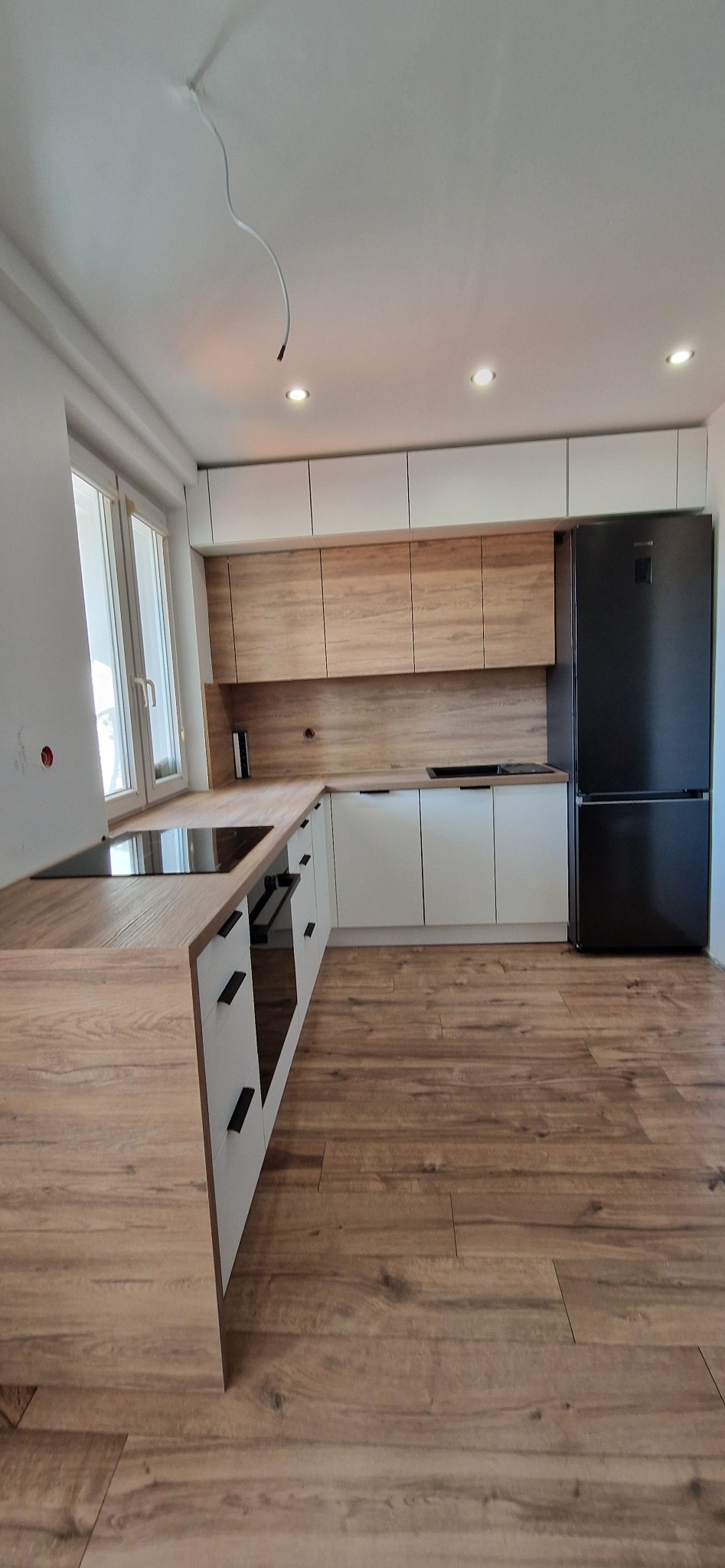 Nowoczesna kuchnia z białymi i drewnopodobnymi frontami, czarnym sprzętem AGD i drewnianą podłogą; widoczny kabel zwisający z sufitu.