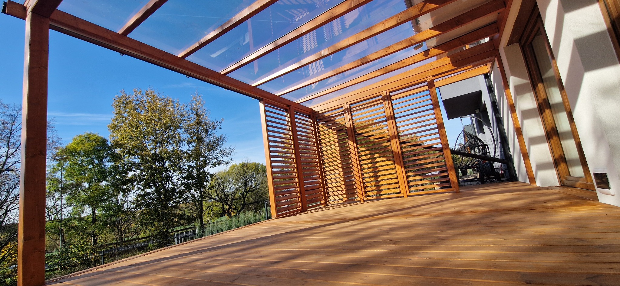 Zadaszony taras z drewnianą podłogą i ażurowymi ścianami, z widokiem na drzewa i błękitne niebo.