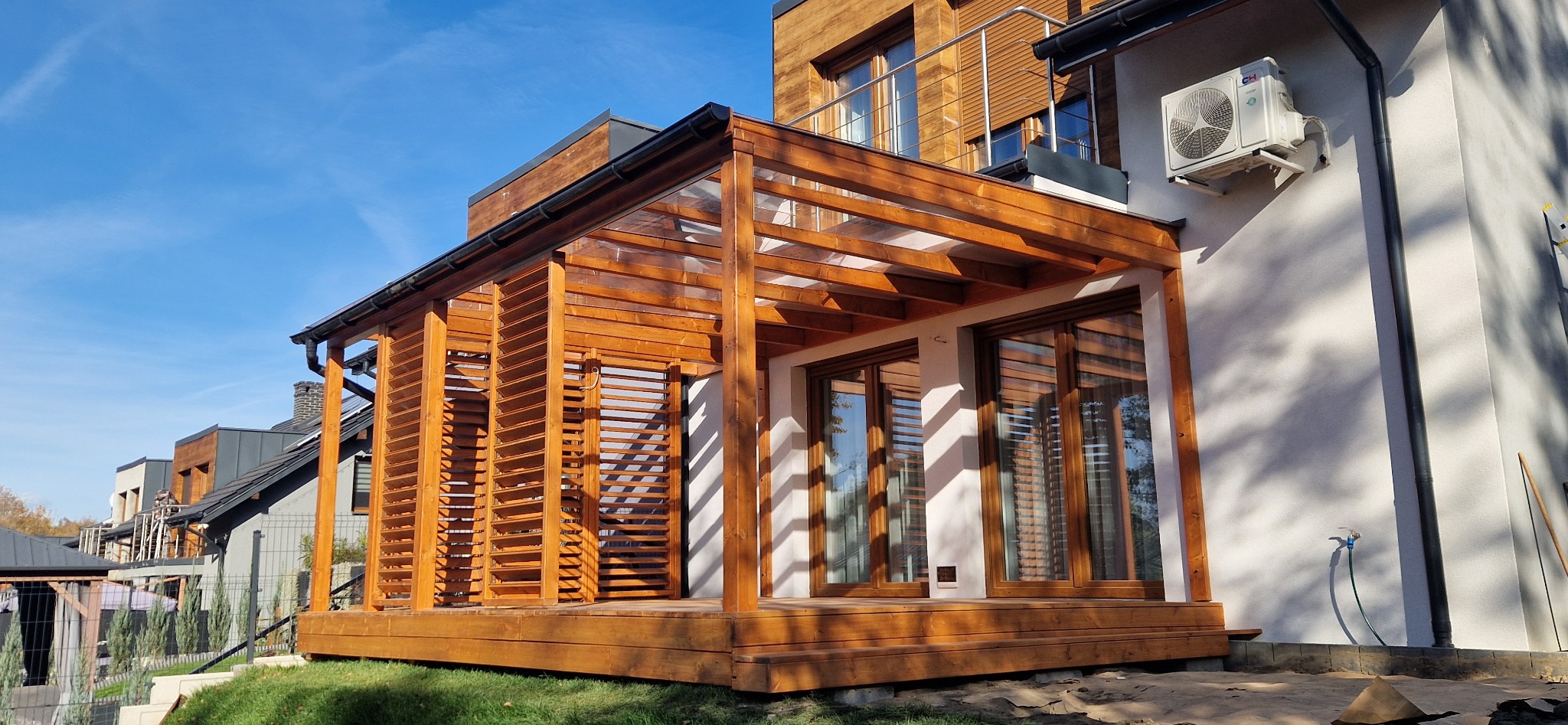Drewniana pergola z lamelami i przezroczystym zadaszeniem przylegająca do białego budynku z klimatyzatorem, widok na błękitne niebo.