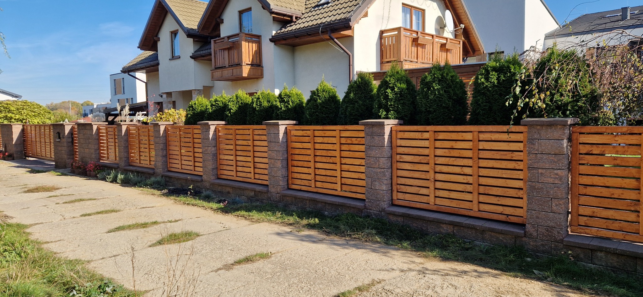 Nowoczesne ogrodzenie z poziomych desek w ciepłym odcieniu drewna, osadzone na kamiennych słupkach i podmurówce, na tle dwupiętrowego domu z balkonami i zadbanym żywopłotem.