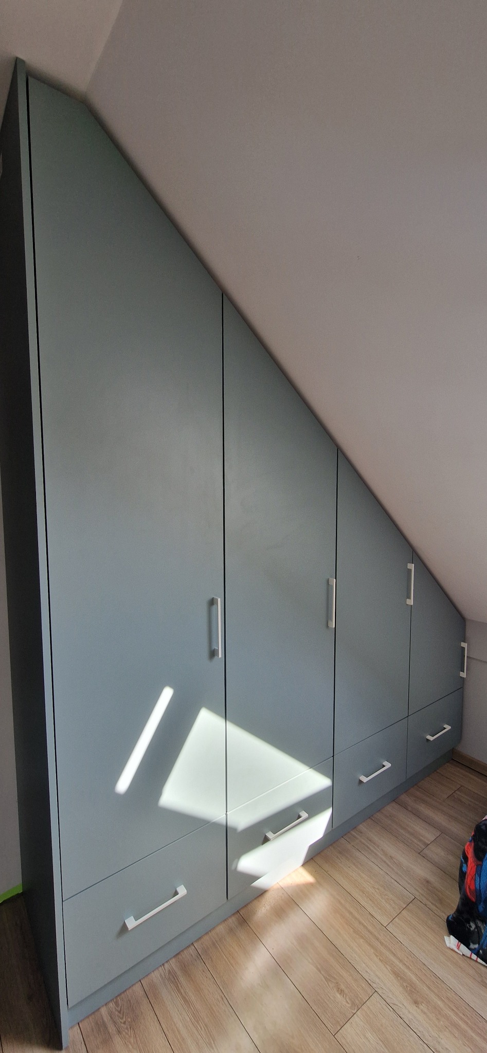 Niebieska zabudowa meblowa ze srebrnymi uchwytami, dopasowana do skosu poddasza, z widocznymi drzwiczkami i szufladami, na podłodze panele.