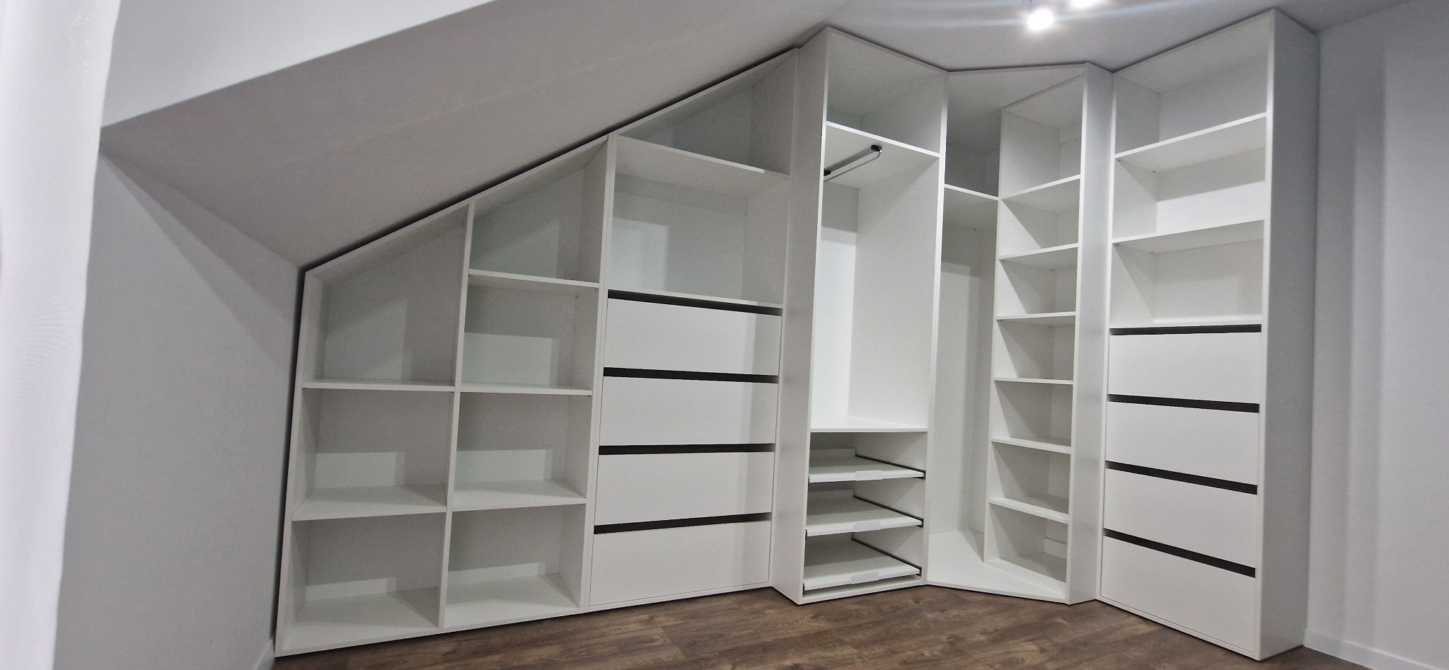 Biała zabudowa meblowa pod skosem dachu, z otwartymi półkami i szufladami z czarnymi akcentami, na drewnianej podłodze.