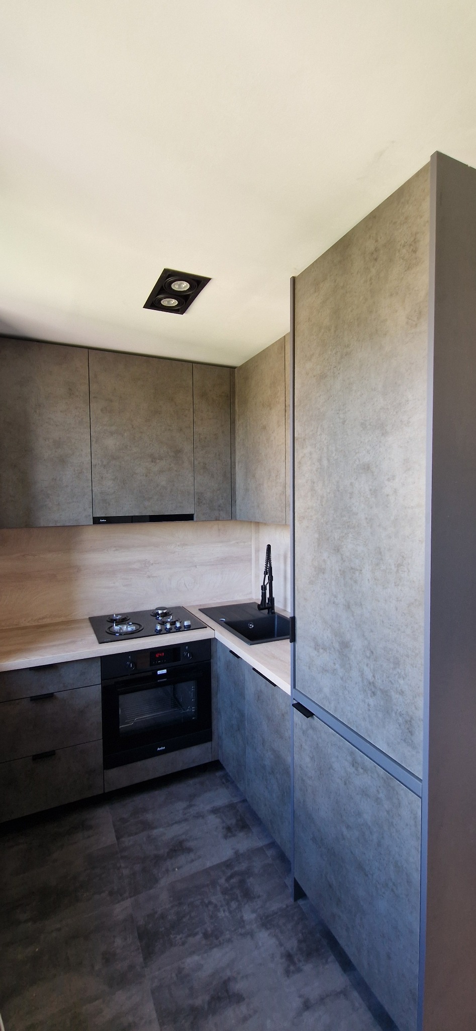 Nowoczesna kuchnia w odcieniach szarości z blatem imitującym drewno, czarnym zlewem i płytą indukcyjną, wykończona matowymi frontami i minimalistycznymi uchwytami.