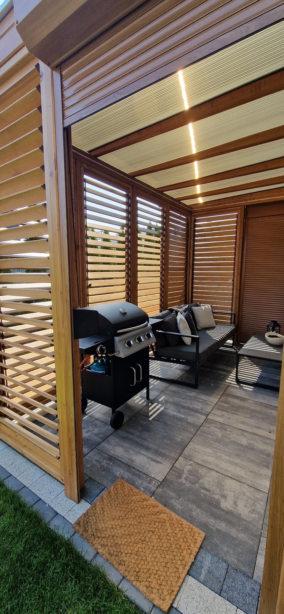 Zadaszony taras z drewnianą konstrukcją żaluzjową w Siemianowicach Śląskich, wyposażony w grill na kółkach i sofę z poduszkami.