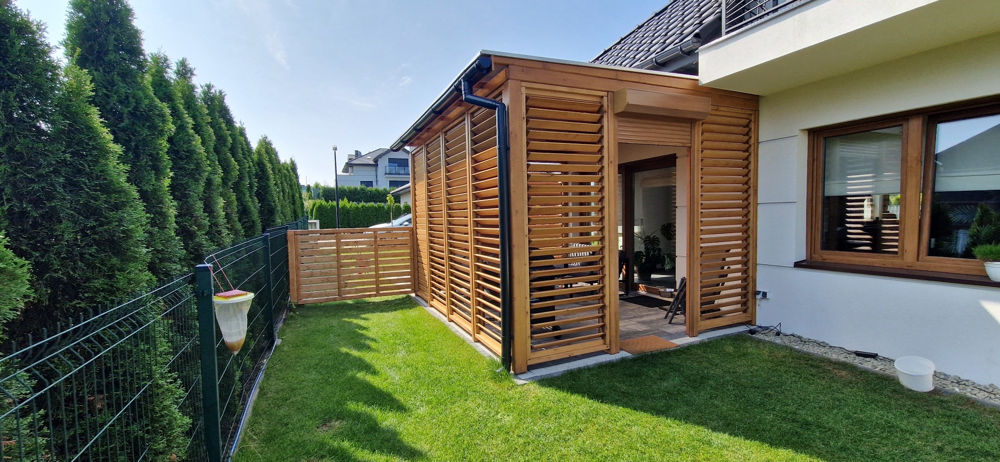 Drewniana konstrukcja z żaluzjowymi ścianami przylegająca do domu z widocznym oknem i zielonym trawnikiem, w tle wysokie tuje i sąsiedni dom.