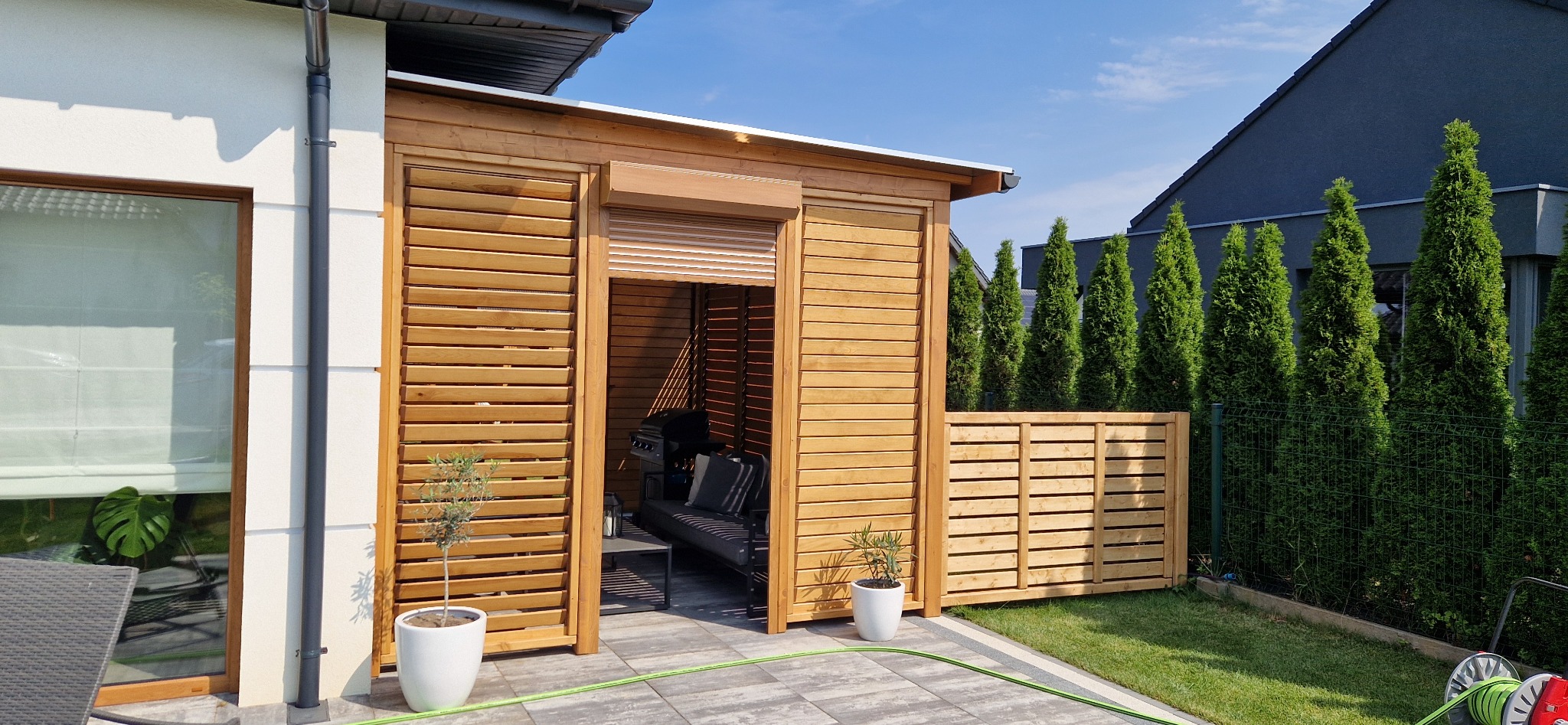 Drewniana pergola z żaluzjami i roletą, wyposażona w sofę i grill, otoczona zielonymi tujami i trawnikiem.