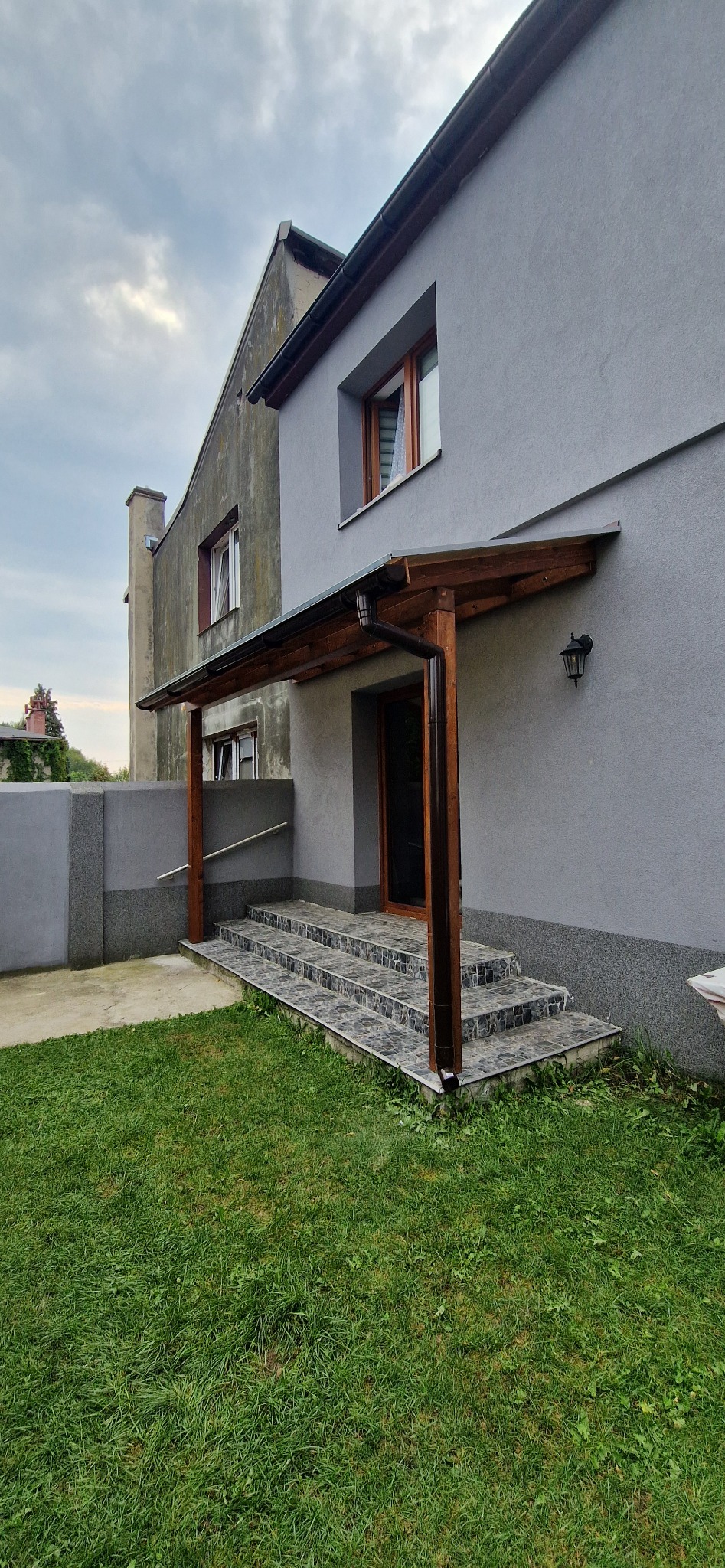 Zadaszone wejście do domu z drewnianą konstrukcją wspartą na słupach, prowadzące po schodach wyłożonych płytkami, z widocznym trawnikiem i fragmentem elewacji budynku.