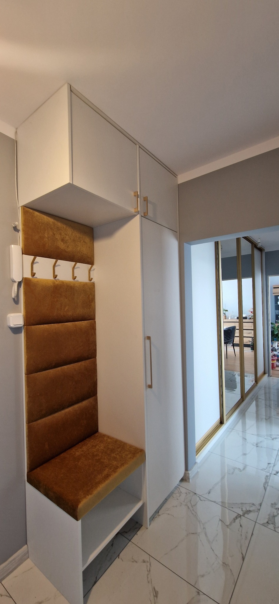 Biała zabudowa przedpokoju z siedziskiem obitym złotym welurem i wieszakami, nad nią szafki, w tle przesuwne drzwi z lustrem.