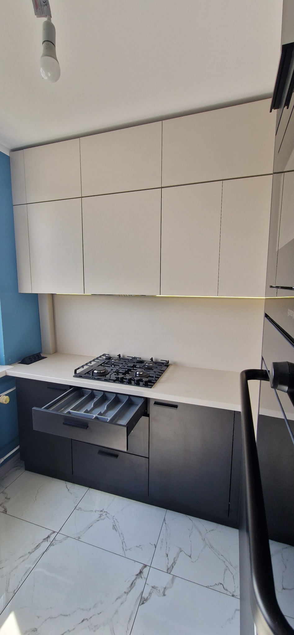 Nowoczesna kuchnia z czarnymi dolnymi szafkami i białymi górnymi, otwartą szufladą na sztućce, płytą gazową i marmurową podłogą.
