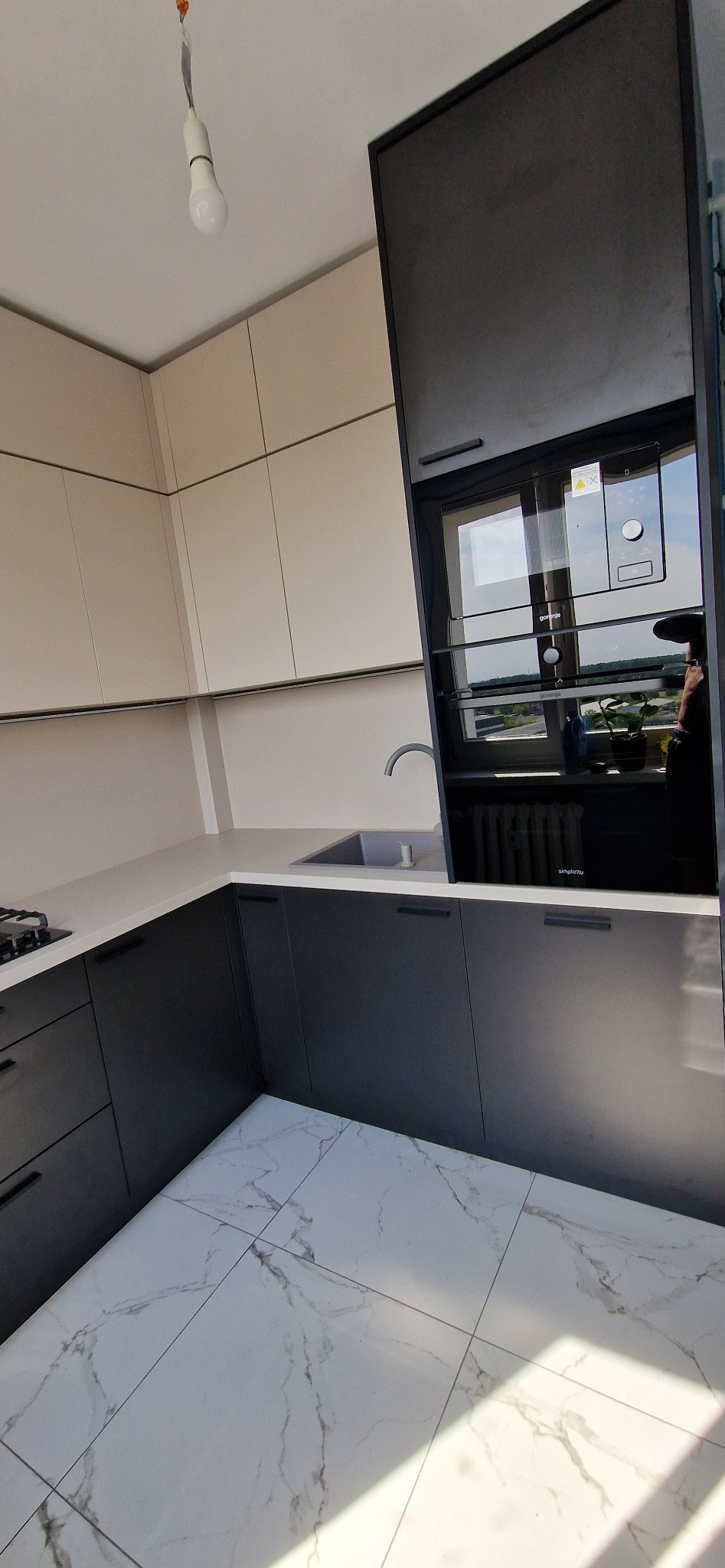 Nowoczesna kuchnia z szarymi szafkami dolnymi i beżowymi górnymi, biały blat, marmurowe płytki podłogowe i widoczne urządzenie AGD.