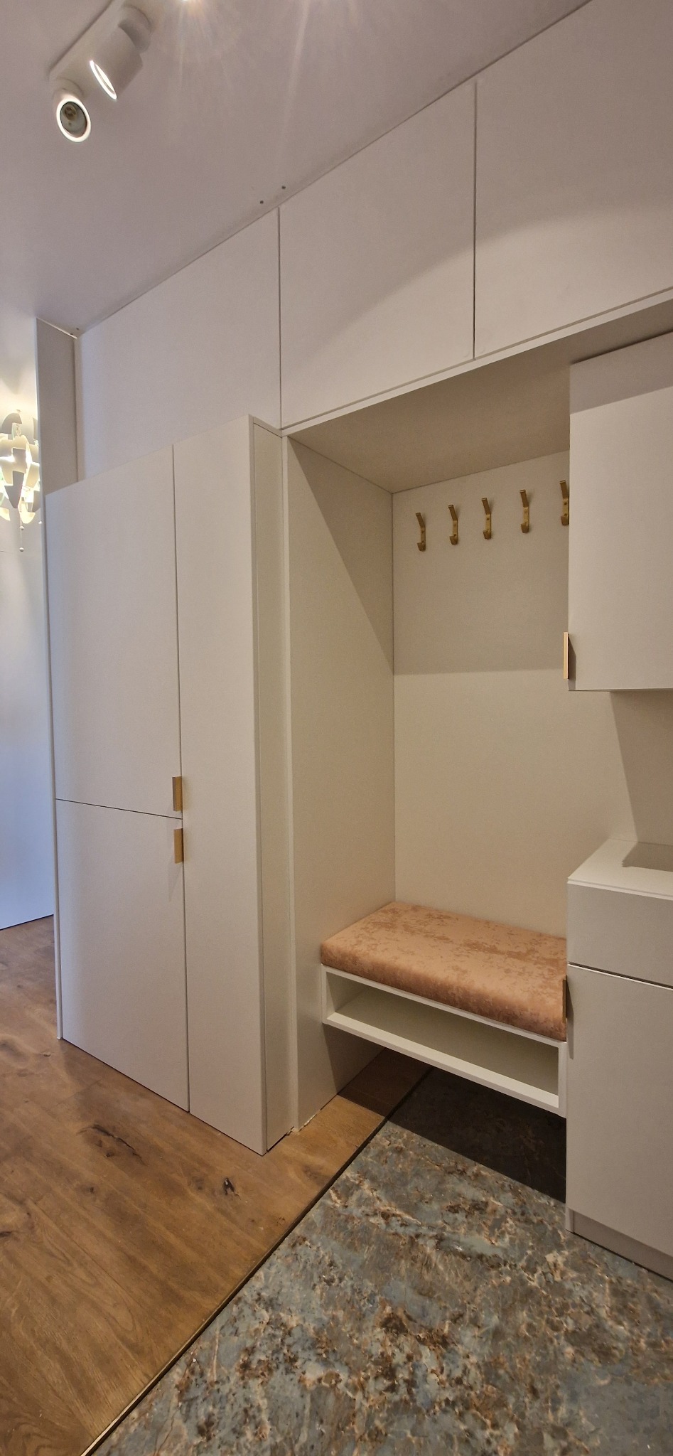 Biała zabudowa przedpokoju z siedziskiem obitym pluszem w odcieniu brzoskwini i złotymi haczykami na ścianie, widoczny fragment drewnianej podłogi oraz podłogi imitującej marmur.