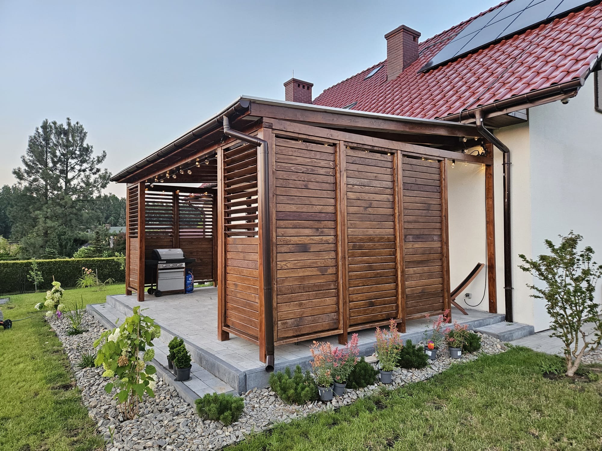 Drewniana konstrukcja z poziomych desek na tarasie, z grillem w środku, oświetlona girlandą żarówek, obok domu z czerwonym dachem i panelami słonecznymi.