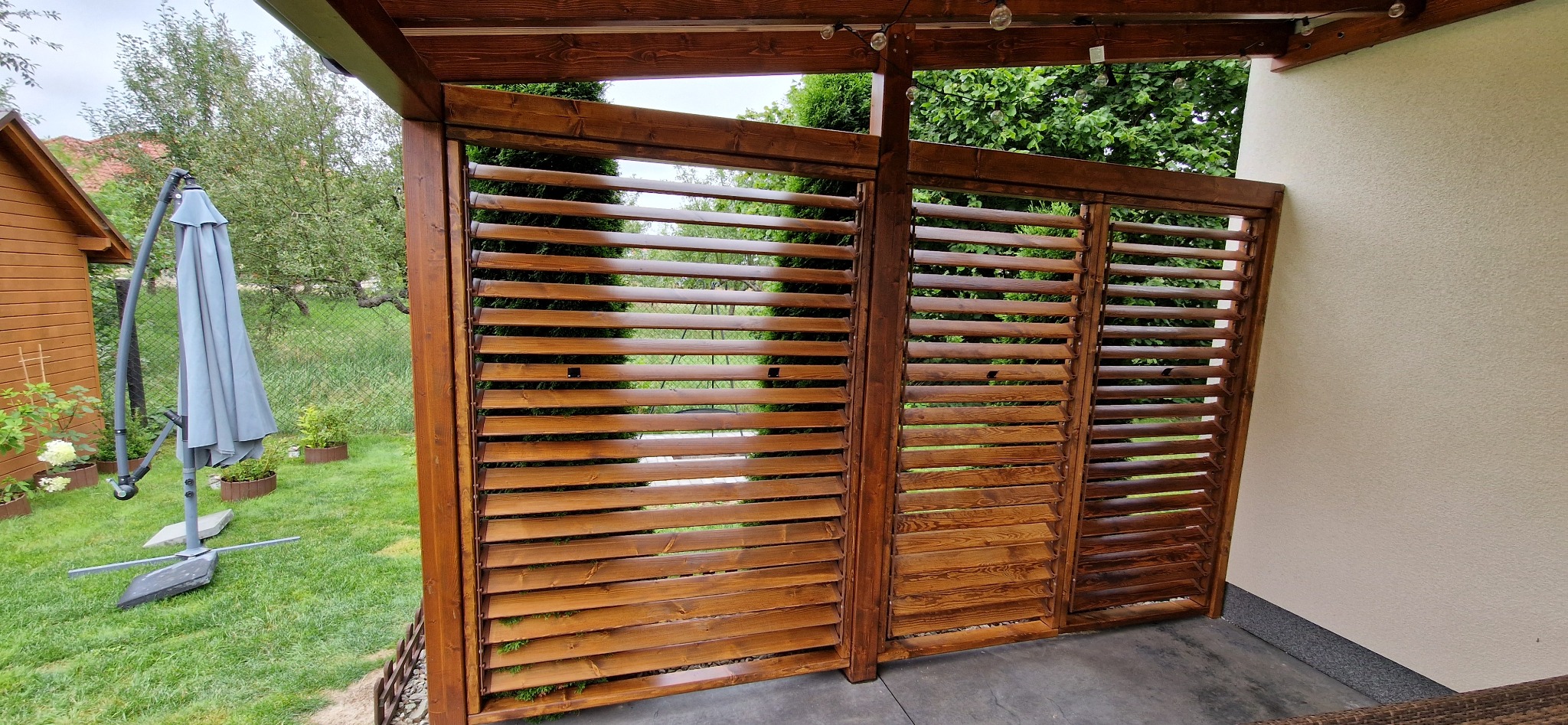 Drewniana pergola z żaluzjami w kolorze ciemnego brązu na tarasie, widok na ogród z parasolem i domkiem narzędziowym.