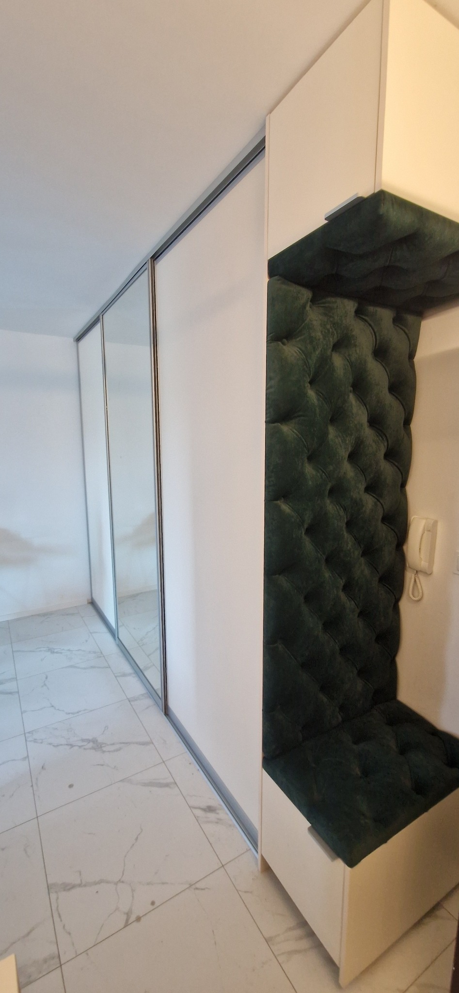 Biała szafa przesuwna z lustrzanymi drzwiami i wbudowanym siedziskiem z ciemnozieloną tapicerką w stylu chesterfield.