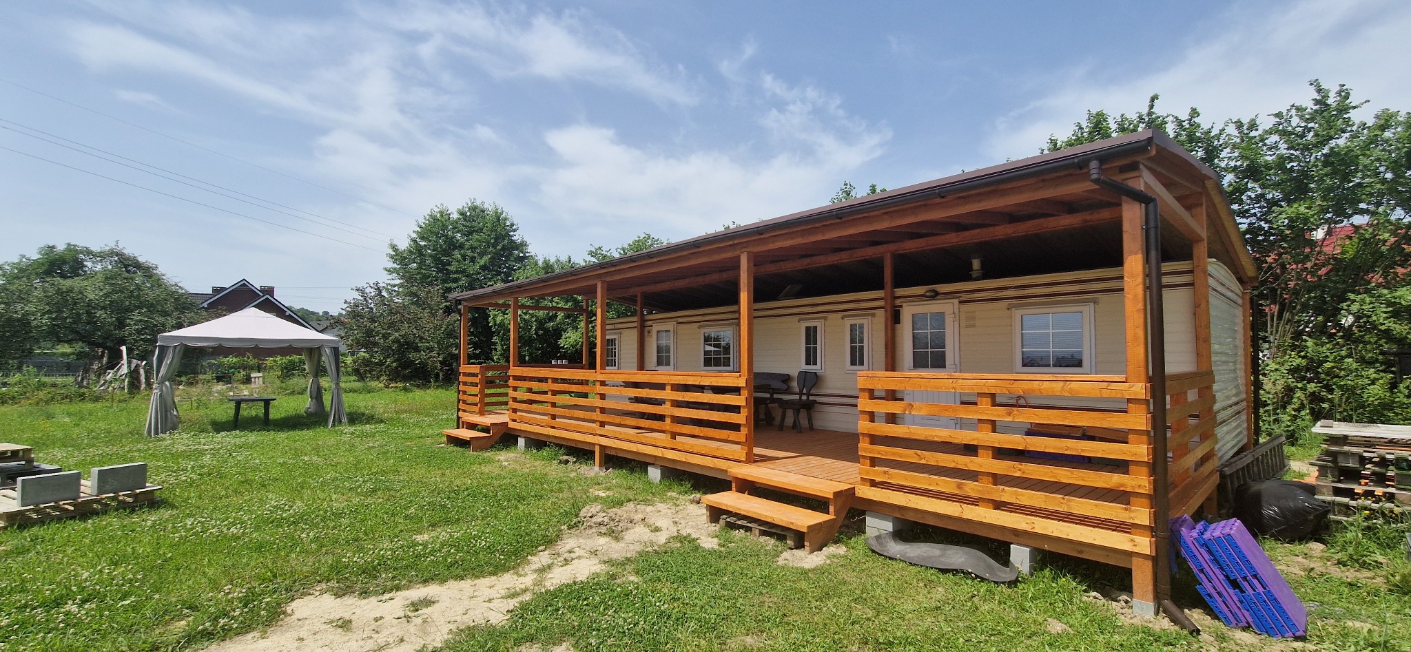 Domek letniskowy z drewnianym zadaszonym tarasem, widok z zewnątrz, w tle biały namiot ogrodowy.
