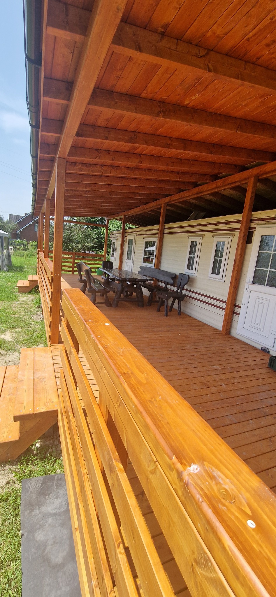 Zadaszony taras z drewnianą podłogą, balustradą i stołem z krzesłami, przylegający do budynku z białymi drzwiami i oknami.