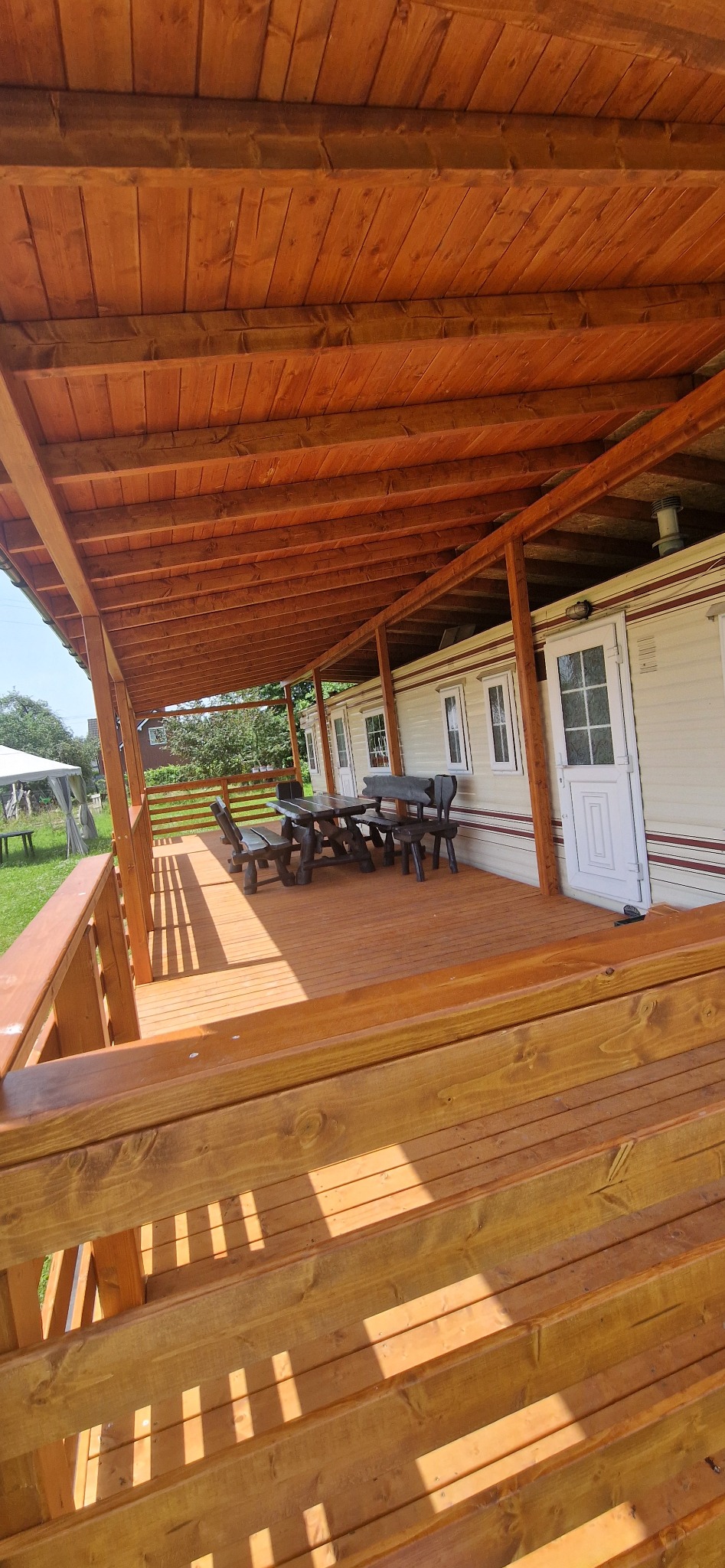 Zadaszony taras z drewnianą podłogą i balustradą, drewniany stół z ławkami i krzesłami, w tle przyczepa kempingowa.