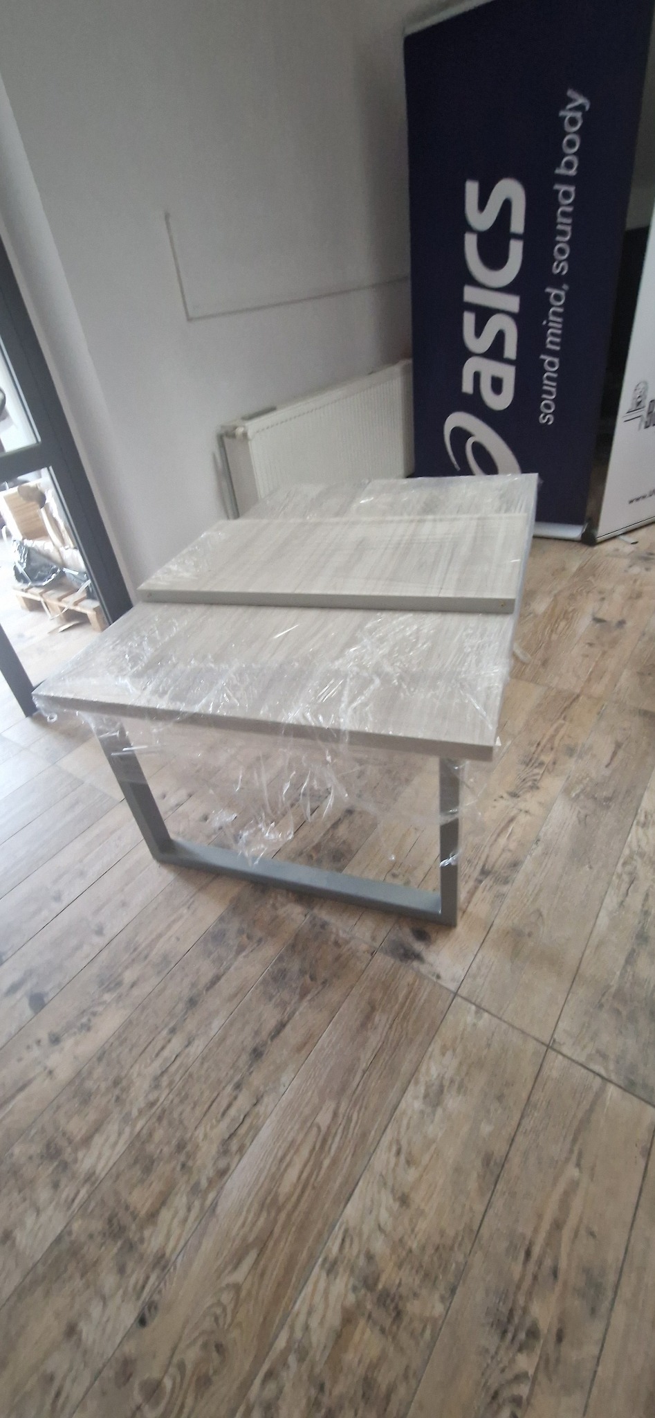 Dwa blaty drewniane zabezpieczone folią, oparte na metalowym stelażu, leżące na podłodze imitującej drewno. W tle grzejnik i baner reklamowy.