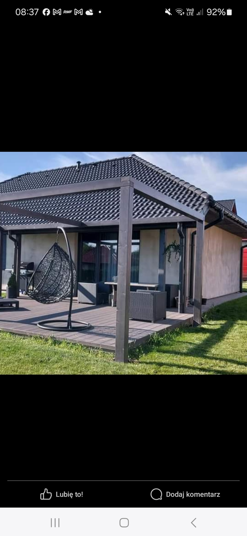 Zadaszony taras z drewnianą konstrukcją, wiszącym fotelem i meblami ogrodowymi na tle domu z ciemną dachówką.