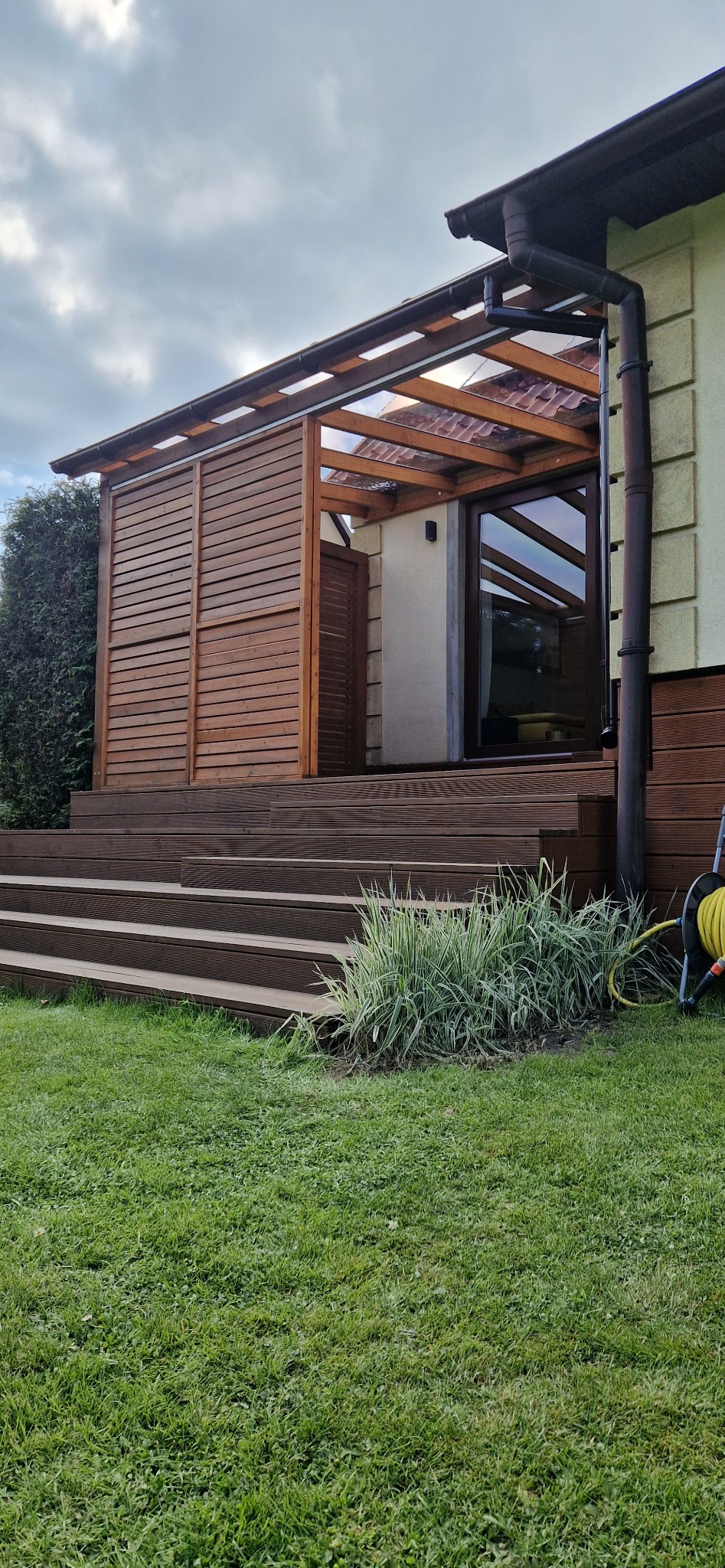 Drewniany taras z zadaszeniem z poliwęglanu, osłonięty panelami z poziomych desek, schody z desek prowadzą na trawnik.