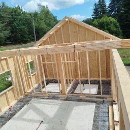 Viktor Kozachuk Uslugi OgulnoBudowlane - Konstrukcja ścian i dachu budynku w trakcie budowy, widok z góry. Drewniane elementy, płyty OSB, narzędzia budowlane. Tło: zielone drzewa i błękitne niebo.