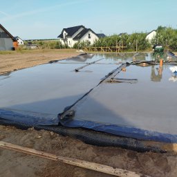 Viktor Kozachuk Uslugi OgulnoBudowlane - Wylana, świeża posadzka fundamentowa zabezpieczona folią budowlaną na gruncie, widoczni pracownicy w oddali. Budowa domu jednorodzinnego w słoneczny dzień.